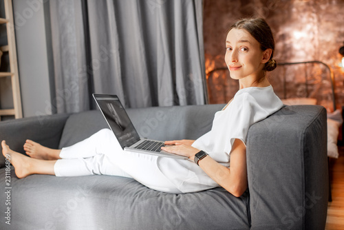 Young woman in white clothes sitting on the couch with laptop at the living room of the modern loft apatment