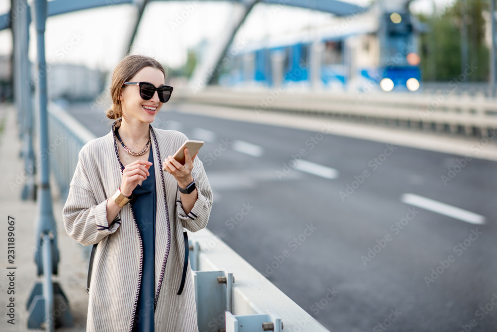 Lifestyle portrait of a young stylish woman with bag and phone outdoors on the modern bridge