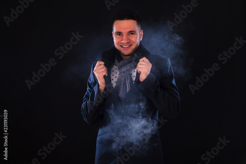 Fototapeta Naklejka Na Ścianę i Meble -  Young male brunette with brown eyes in the studio on a black background and white smoke, dressed in a dark gray coat, checkered scarf. Cold weather concept, steam from mouth, beginning of winter