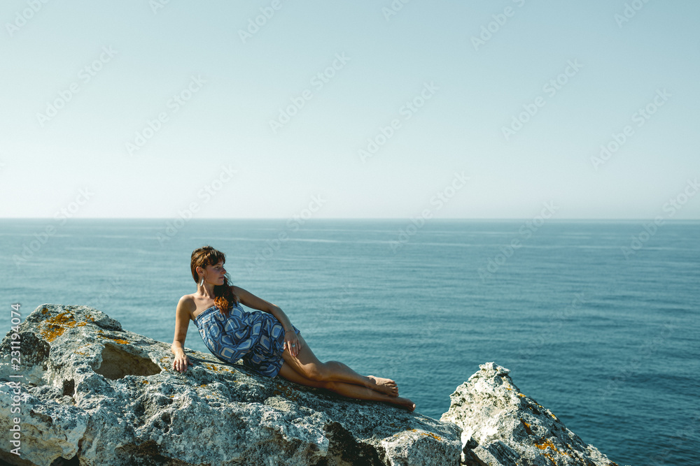 The concept of freedom and relaxation. A young woman with slender tanned legs sits on a rock on a cliff near a blue, deep sea. Place to advertise.