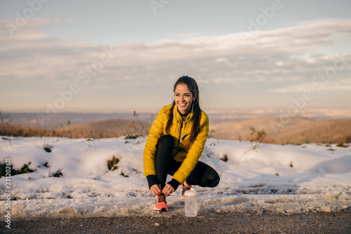 Happy sporty woman crouching and tying shoelace on the road in. In front of her bottle with water, wintertime.