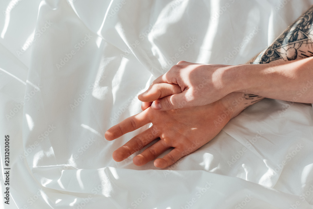 Fototapeta premium partial view of loving couple holding hands while lying on white bed sheet