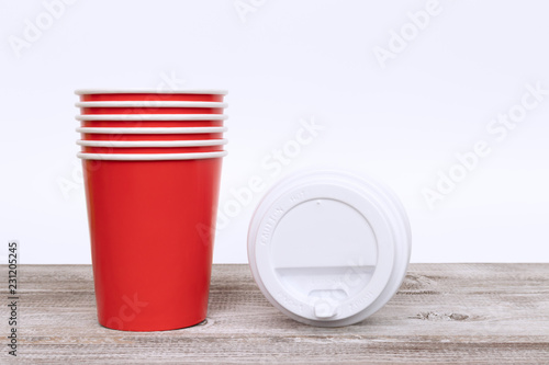 A stack of red take away disposable paper cups and plastic caps on wooden desk and white background