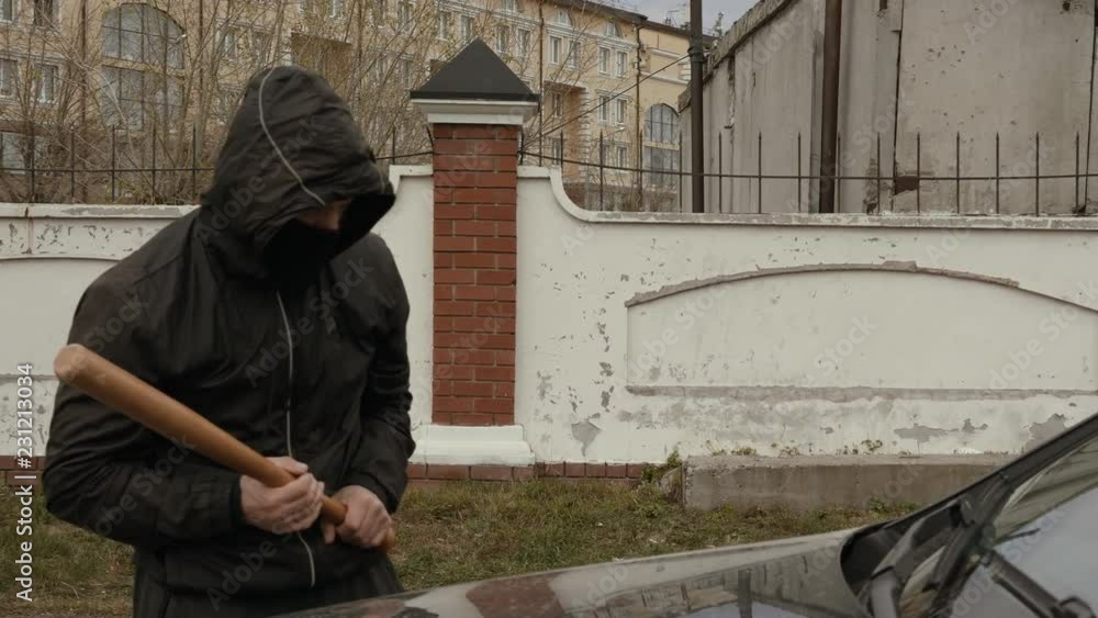 Aggressive hooligan in mask and hood crashing car with baseball bat Aggressive hooligan in mask and hood crashing car with baseball bat