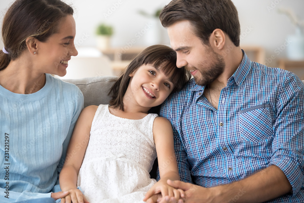 Attractive positive family with little daughter sitting together on sofa at home. Adorable girl smiling looking at camera loving mother and father holds her hands. Happy multi-ethnic family concept Attractive positive family with little daughter sitting together on sofa at home. Adorable girl smiling looking at camera loving mother and father holds her hands. Happy multi-ethnic family concept