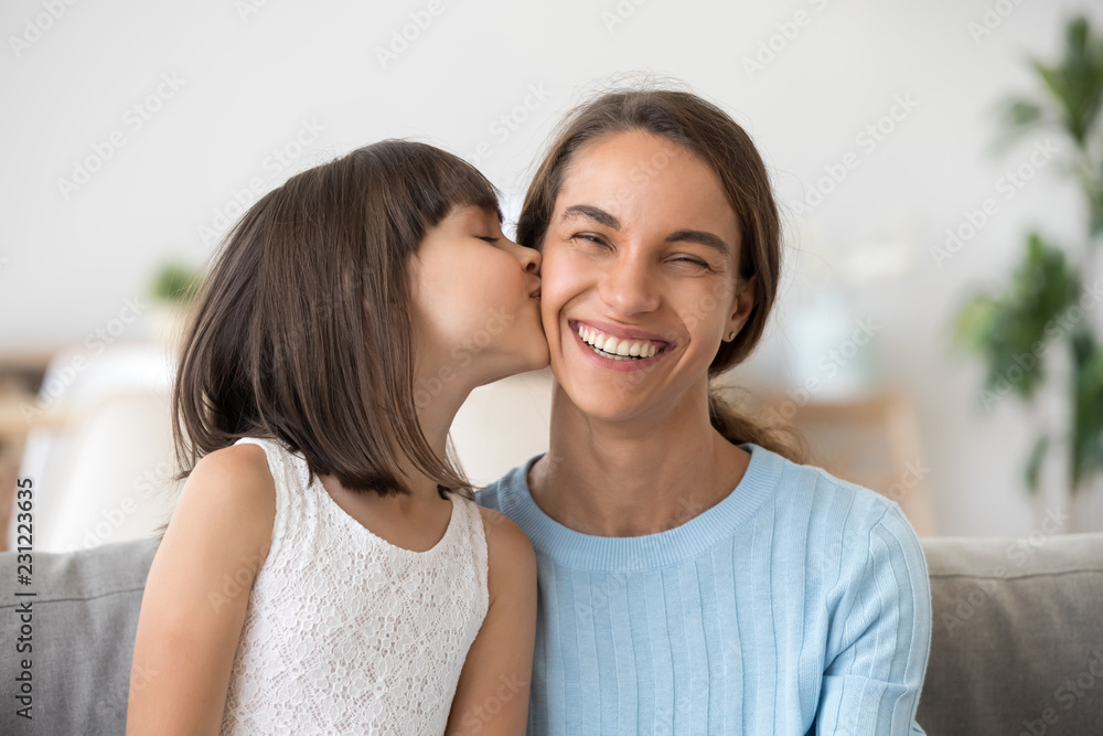 Head shot portrait smiling mother smiling looking at camera her daughter kisses her on cheek sitting together on couch at home. Happy motherhood warm relations and new mum for adopted child concept Head shot portrait smiling mother smiling looking at camera her daughter kisses her on cheek sitting together on couch at home. Happy motherhood warm relations and new mum for adopted child concept