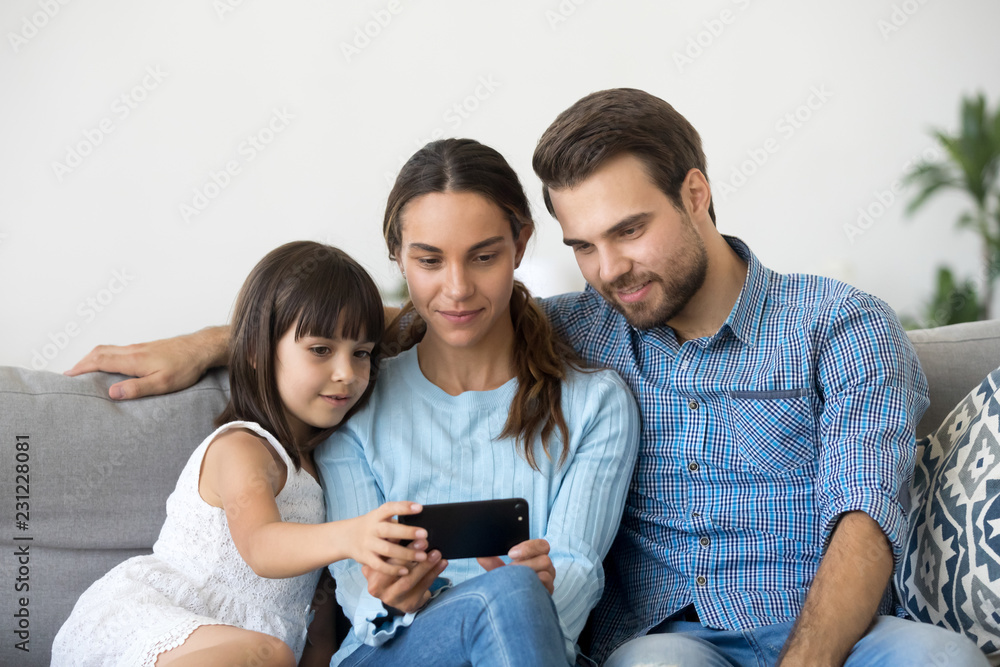Married diverse couple spend free time with little daughter sitting together on couch in living room at home. Positive mother father small child using mobile phone watching cartoons play games online Married diverse couple spend free time with little daughter sitting together on couch in living room at home. Positive mother father small child using mobile phone watching cartoons play games online