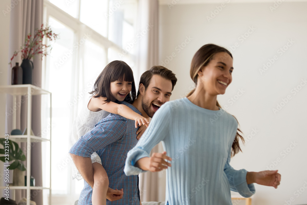 Young happy father laughing carrying on back piggybacking little daughter catching mum playing with diverse family having fun together. Kid enjoying active game with parents in living room at home Young happy father laughing carrying on back piggybacking little daughter catching mum playing with diverse family having fun together. Kid enjoying active game with parents in living room at home
