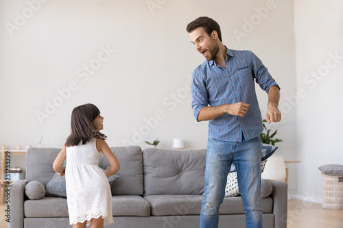 Cheerful young father and little daughter standing in living room at home moving dancing to favourite song together. Child having fun with loving daddy. Active lifestyle leisure activities concept