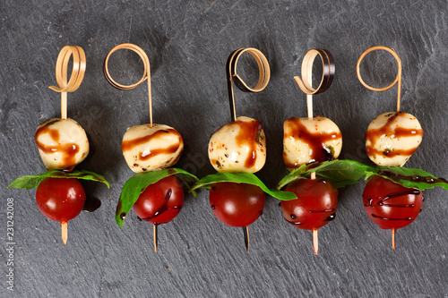 Caprese salad skewers with mozzarella, basil, tomatoes and balsamic glaze. Appetizers against a dark stone background.