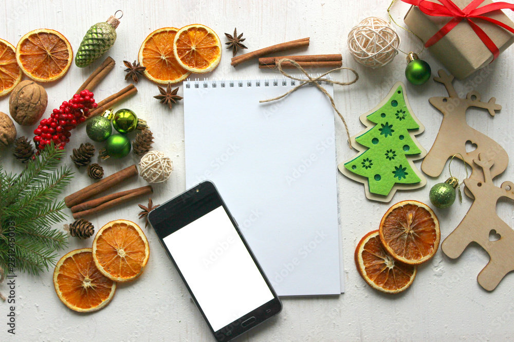 Fototapeta premium Christmas card, blank sheet of paper and mobile phone surrounded by Christmas balls, fir branches, cinnamon sticks, spices, dried orange, wooden deer 