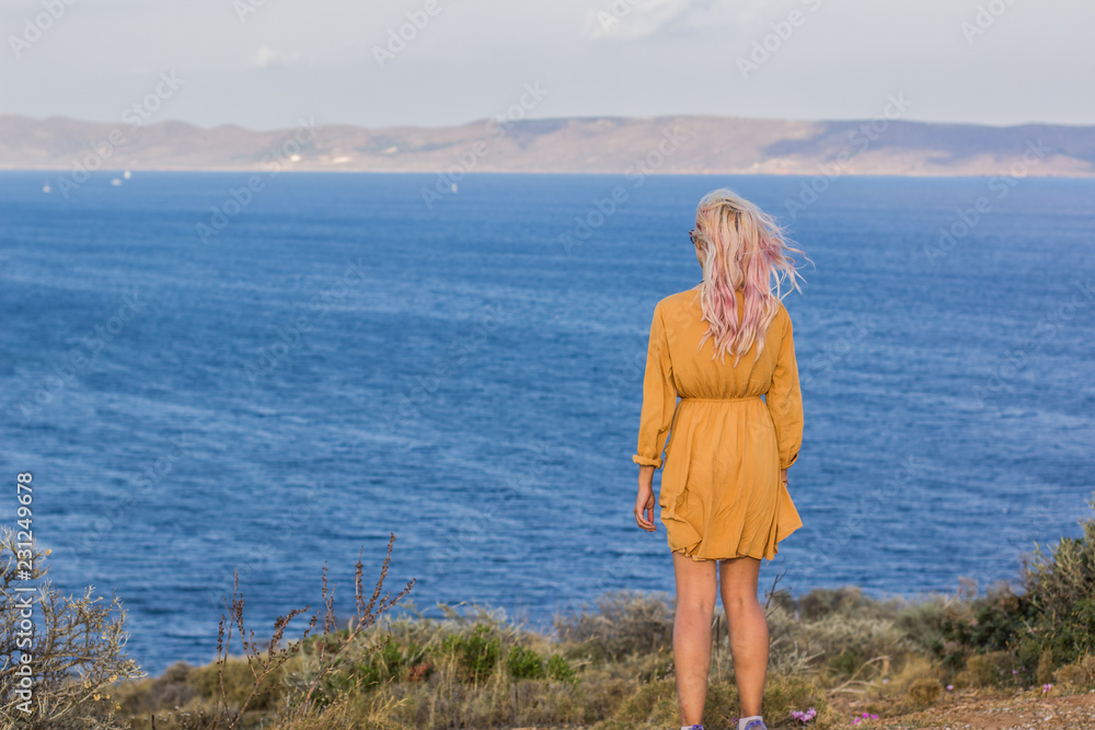 freedom lifestyle travel and adventure concept of young vagabond girl in yellow dress stay back to camera on beautiful south top of cape mountain above blue sea water surface in summer weather time