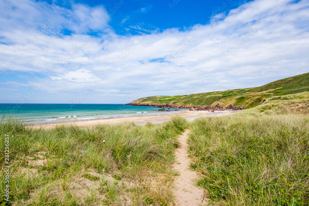 Scenic landscape of Pembrokeshire coast, Uk.Path on sand dune covered with grass patches leading to paradise beach with turquoise sea.Bright and vibrant landscape image with copy space. Scenic landscape of Pembrokeshire coast, Uk.Path on sand dune covered with grass patches leading to paradise beach with turquoise sea.Bright and vibrant landscape image with copy space.