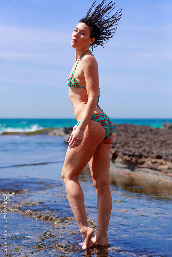 Striking young woman green red bikini flicking wet hair back on rock  platform ocean behind Stock Photo | Adobe Stock