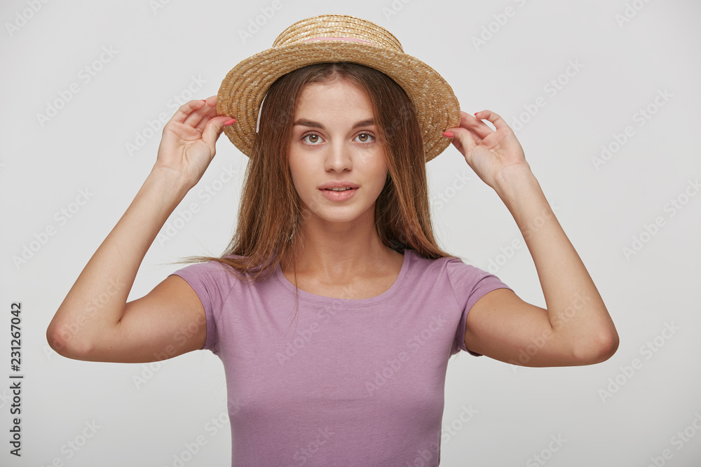 Charming young female keeps in hands straw hat, tries it on or corrects, imagines a vacation by the sea, hot summer, dreams of summer trip, against white studio background Charming young female keeps in hands straw hat, tries it on or corrects, imagines a vacation by the sea, hot summer, dreams of summer trip, against white studio background