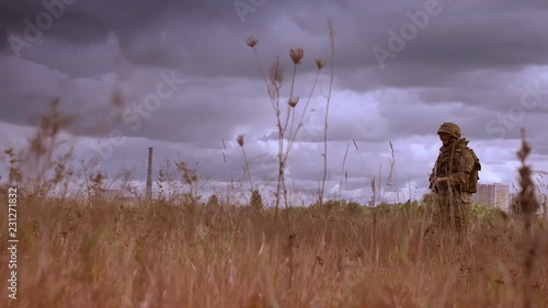 Resistant strong soldier holding automatic weapon and crossing wheat field alone in camouflage, dark cloudy sky during daytime, authentic view