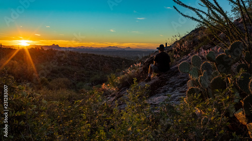 A man in a broad brimmed hat, t-shirt and shorts with hiking boots, sitting on a rock in the Sonoran Desert watching the sunset. Linda Vista trail, Oro Valley, Arizona, USA. © Charles