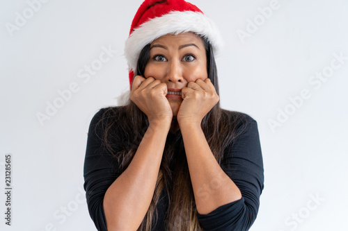 Scared woman in Christmas hat falling into panic. Nervous mix raced lady covering cheeks with fists. Panic concept