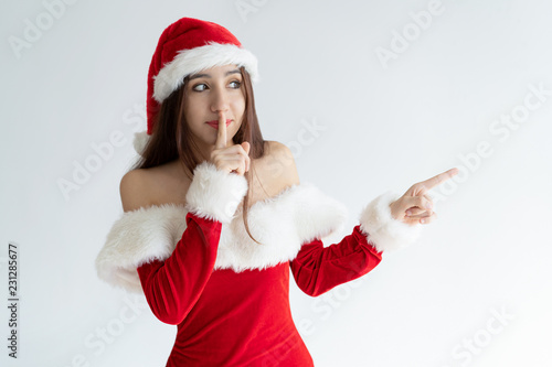 Portrait of calm woman standing with finger on lips and pointing at something. Young mixed race girl wearing Christmas outfit gossiping. Christmas or New Year sale concept