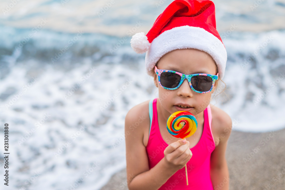 Obraz premium Christmas background: cute child in Santa hat celebrating New Year and Christmas on the beach, free space.
