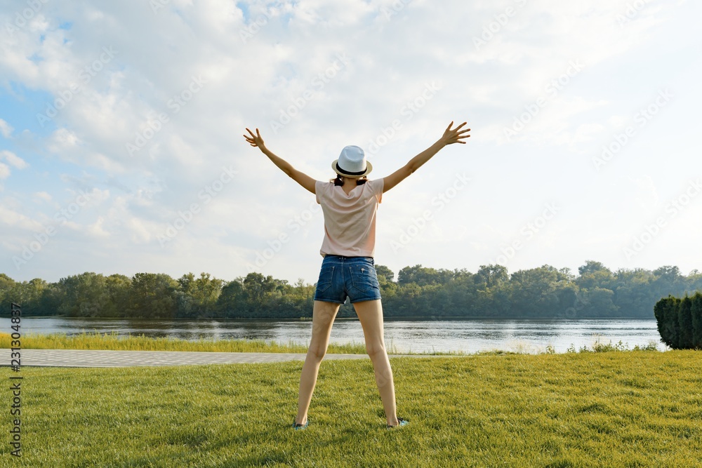 Young girl with her back in hat on the river bank, spread her hands in the sides, looks at the setting sun. Enjoys and meditates Young girl with her back in hat on the river bank, spread her hands in the sides, looks at the setting sun. Enjoys and meditates