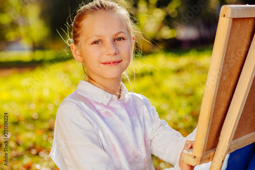 Cute little girl school holding brush painting colorful picture with easel outdoor in park. Open air activity for school age children concept.