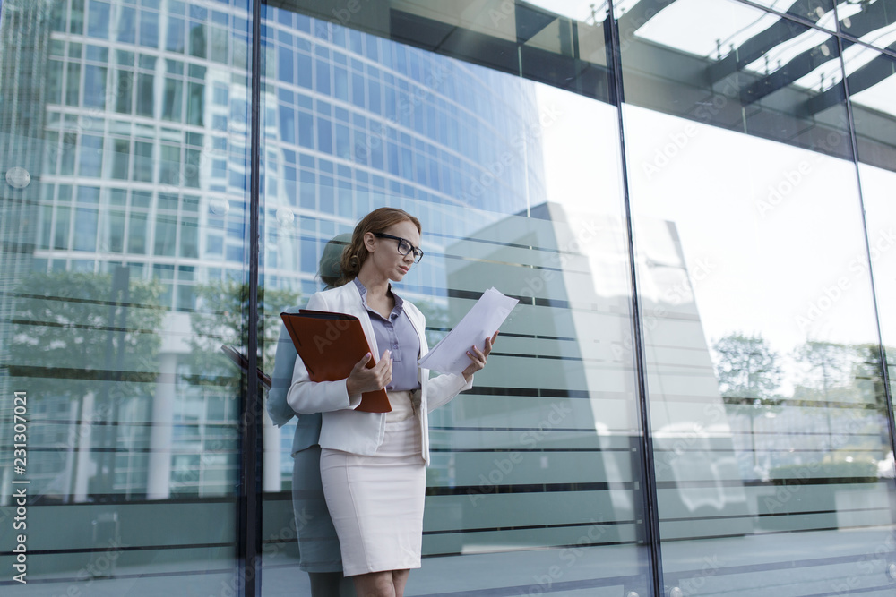 The young business woman in gray office dress holds in hand folder with documents. Businesswoman standing next to the business center
