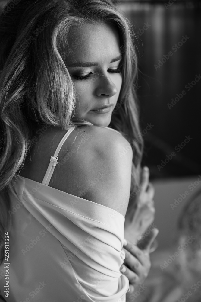 Woman portrait at party. Lovely, beautiful, young girl in gentle silk pajama, holding a glass of champagne, posing while sitting on the bed. Indoors. Black and white photo. Woman portrait at party. Lovely, beautiful, young girl in gentle silk pajama, holding a glass of champagne, posing while sitting on the bed. Indoors. Black and white photo.