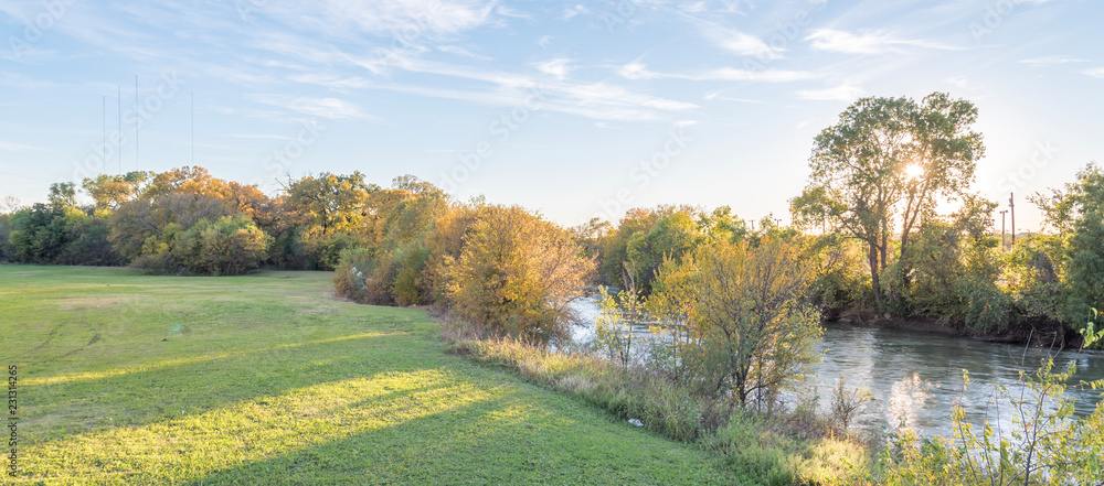 Naklejka premium Panorama view beautiful riverside park with fall foliage vibrant color and sun peeking through trees in Carrollton, Texas, USA. Stunning yellow autumn leaves at sunset