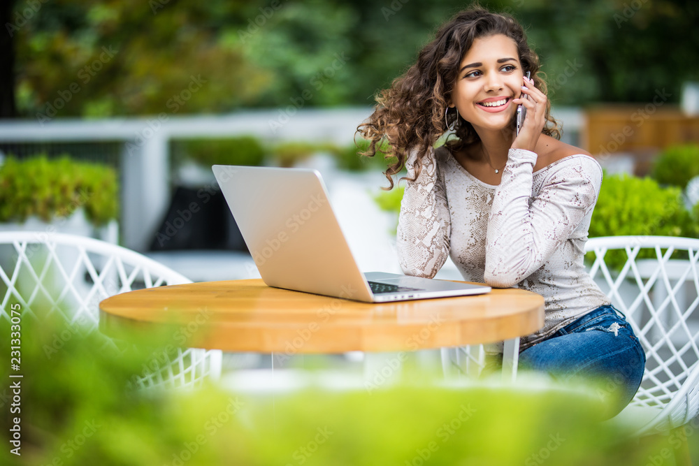 Young curly latin woman calling with cell telephone while sitting alone in street cafe during free time while work on her modern laptop Young curly latin woman calling with cell telephone while sitting alone in street cafe during free time while work on her modern laptop