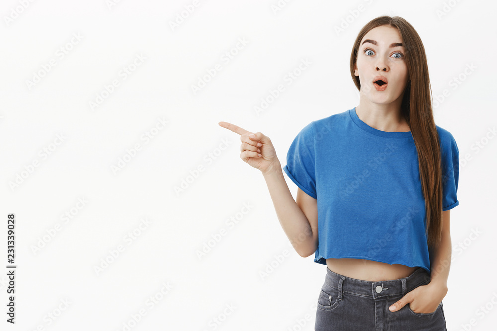 Amused and thrilled enthusiastic attractive brunette in stylish blue t-shirt gasping folding lips from amazement pointing left being astonished with awesome copy space against white background Amused and thrilled enthusiastic attractive brunette in stylish blue t-shirt gasping folding lips from amazement pointing left being astonished with awesome copy space against white background