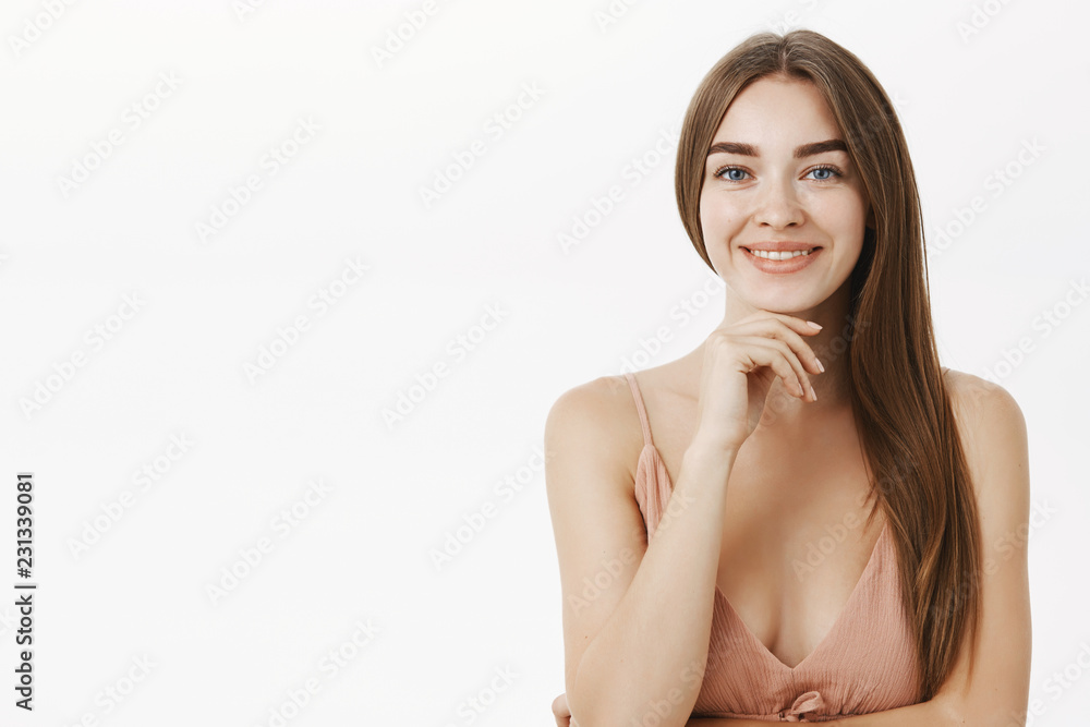 Fototapeta premium Waist-up shot of sensual and feminine brunette in cute beige dress holding hand on chin and smiling broadly having pure clear skin delighted to have no problems using minimal makeup over grey wall