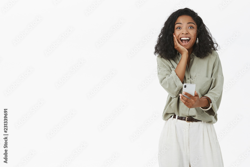 Girlfriend receive first payment on online bank account rejoicing feeling happy and surprised touching cheek looking with joy and amazement at camera holding smartphone posing against gray background Girlfriend receive first payment on online bank account rejoicing feeling happy and surprised touching cheek looking with joy and amazement at camera holding smartphone posing against gray background
