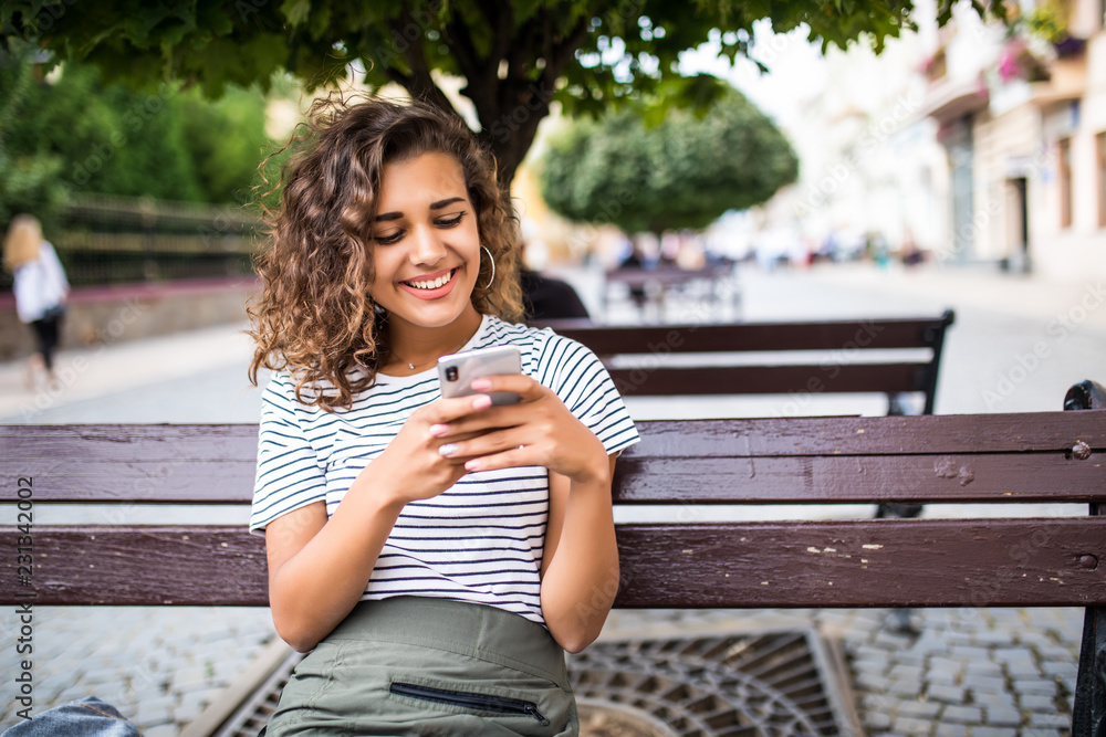 Pretty mixed race girl with curly hair use smartphone while sitting on the bench outdoor Pretty mixed race girl with curly hair use smartphone while sitting on the bench outdoor