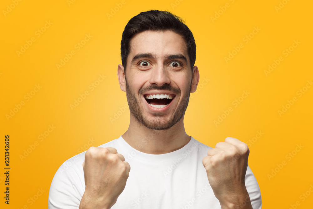 Young man shouting while his team win, raised both fist in victory gesture, isolated on yellow background Young man shouting while his team win, raised both fist in victory gesture, isolated on yellow background
