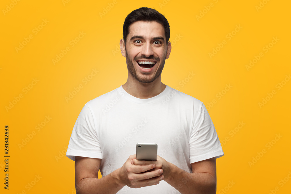 Portrait of handsome young man in white t-shirt, holding smartphone and laughing, isolated on yellow background Portrait of handsome young man in white t-shirt, holding smartphone and laughing, isolated on yellow background