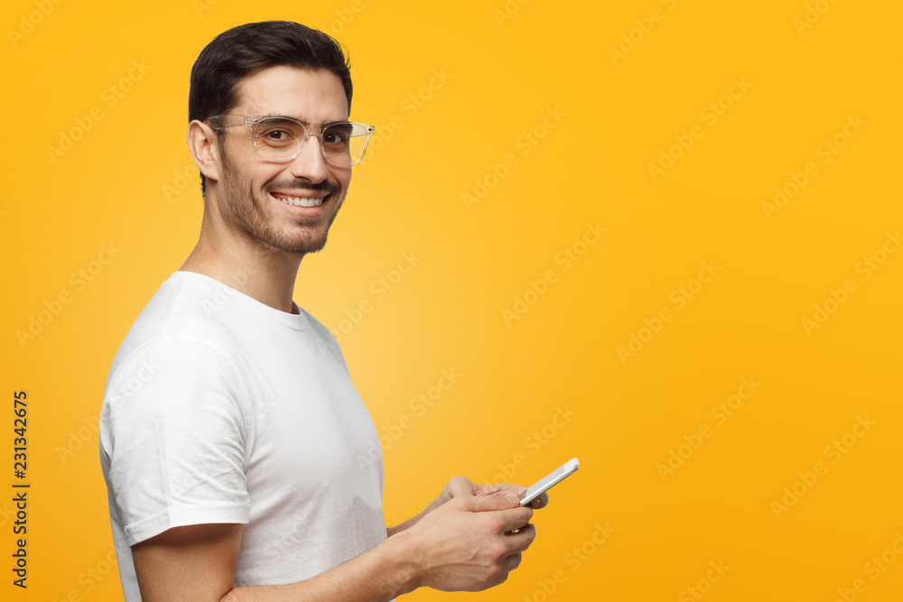 Side portrait of young handsome caucasian man isolated on yellow background, holding smartphone in hands with open positive smile Side portrait of young handsome caucasian man isolated on yellow background, holding smartphone in hands with open positive smile