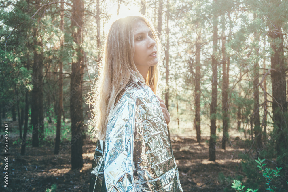 Beautiful girl is wrapped in a foil emergence blanket. Wet model stands in the forest in the morning. Concept of the tourist who studied the problems of pollution of nature who was wet from the water