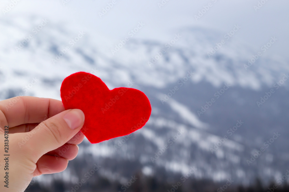 Fototapeta premium Hand holding red felt heart on the winter mountains landscape snow covered background. Love winter, ski concept. Copy space