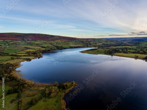Fototapeta Naklejka Na Ścianę i Meble -  Aerial panoramic view of a beautiful natural lake in Brecon Beacons surrounded by rural farmland (Llangorse Lake, Wales)