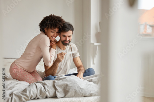 Parenthood, good news, baby expecting concept. Joyful dark skinned wife leans shoulder of her husband, look positively at pregnancy test, going to have child. Family couple on bed in modern apartment photo