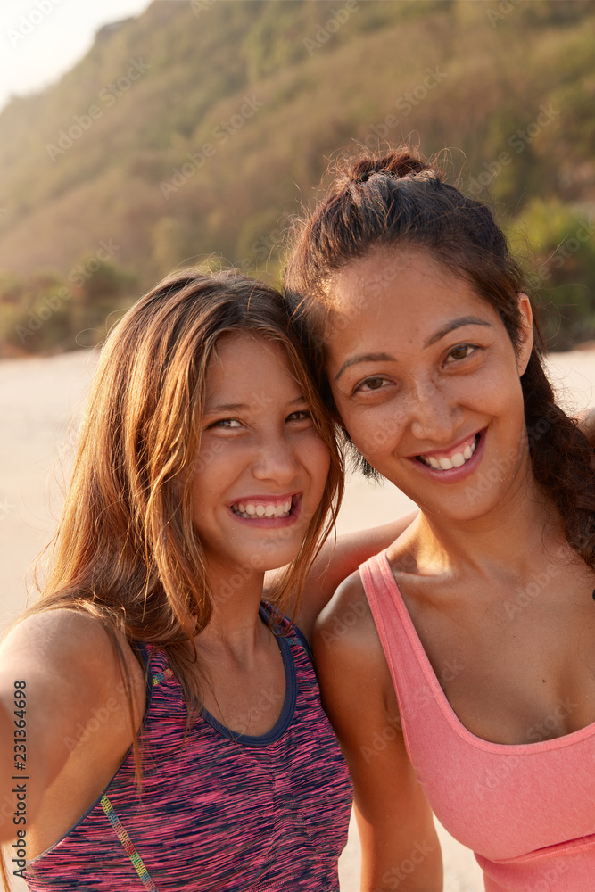Smiling mixed race female friends pose near for making selfie, dressed in top, have leisure time for sport, model against cliff background, enjoy summer holidays. Friendship, relationships concept Smiling mixed race female friends pose near for making selfie, dressed in top, have leisure time for sport, model against cliff background, enjoy summer holidays. Friendship, relationships concept