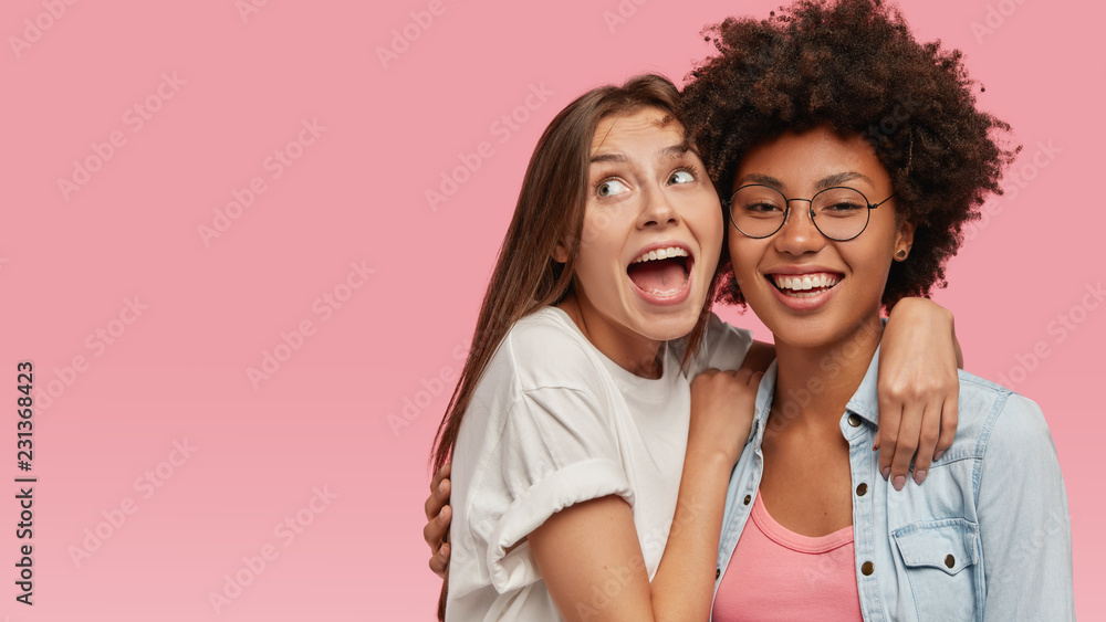 Overjoyed young European woman with widely opened mouth, has joy and embraces her older Afro American sister, enjoy pleasant moments, laugh at something funny, model in pink studio with blank space Overjoyed young European woman with widely opened mouth, has joy and embraces her older Afro American sister, enjoy pleasant moments, laugh at something funny, model in pink studio with blank space