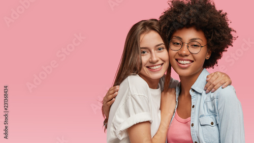 Multiracial cheerful young best friends embrace affectionately, have toothy smiles, stand closely to each other, isolated against pink background with free space for your promotional content photo