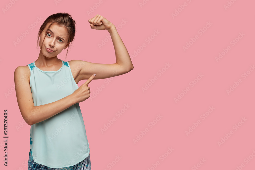 Healthy lifestyle and sport concept. Dissatisfied European woman points at arm muscle, wants to have muscular body, poses over pink background with free space for your advertisement or promotion Healthy lifestyle and sport concept. Dissatisfied European woman points at arm muscle, wants to have muscular body, poses over pink background with free space for your advertisement or promotion