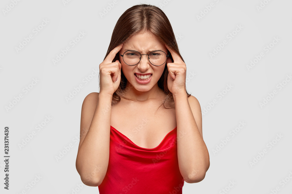 Stressed brunette lady touches temples and thinks hard, has terrible headache, frowns face in displeasure, wears red elegant dress, needs rest, poses over white background. Negative feelings concept Stressed brunette lady touches temples and thinks hard, has terrible headache, frowns face in displeasure, wears red elegant dress, needs rest, poses over white background. Negative feelings concept