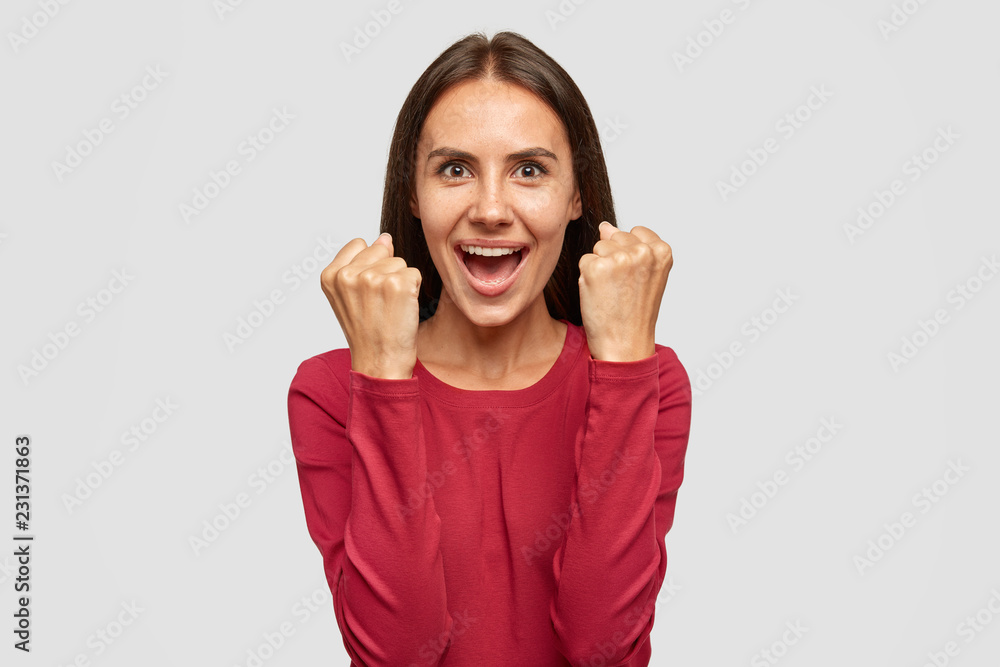 Waist up shot of European brunette girl clenches fists with triumph, dressed in red casual sweater, celebrates successful deal with business partner, models against white background. Body language Waist up shot of European brunette girl clenches fists with triumph, dressed in red casual sweater, celebrates successful deal with business partner, models against white background. Body language