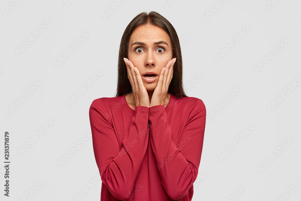 Stupefied woman with black hair, gazes with dark eyes, keeps hands on cheeks, dressed in red sweater, poses against white studio background, gasps from amazement. People and emotions concept