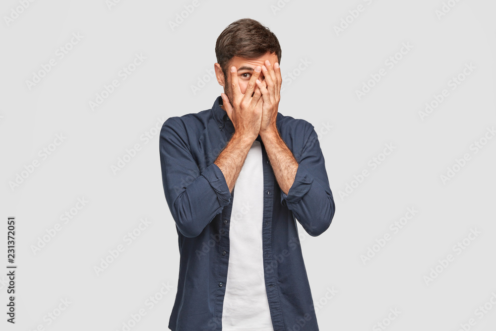 Handsome man with dark hair, bristle, hides face with hands, peeks through fingers, wears fashionable shirt, feels shy, isolated over white background. Curious guy cannot to look at present. Handsome man with dark hair, bristle, hides face with hands, peeks through fingers, wears fashionable shirt, feels shy, isolated over white background. Curious guy cannot to look at present.