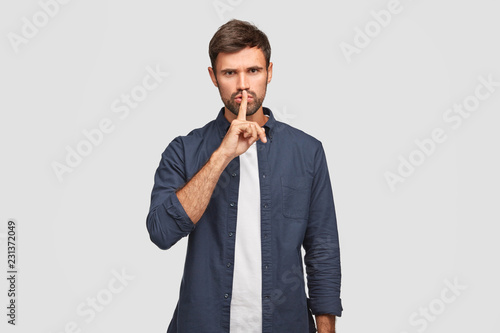 Photo of serious male keeps index finger on lips as demonstrates hush gesture, asks be quiet, dressed in fashionable clothes, angry as someone tells his secret, isolated over white background